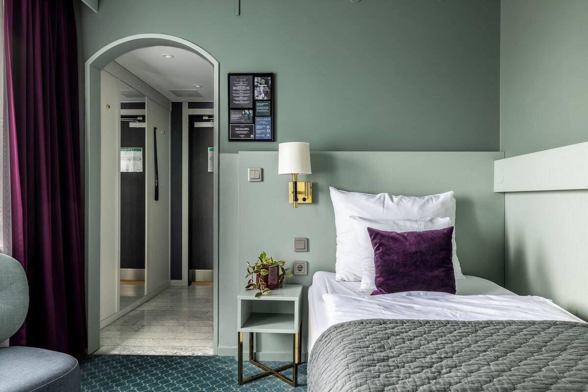 Une chambre d’hôtel avec un lit, une table de chevet, une lampe, une plante en pot et un panneau fixé au mur.