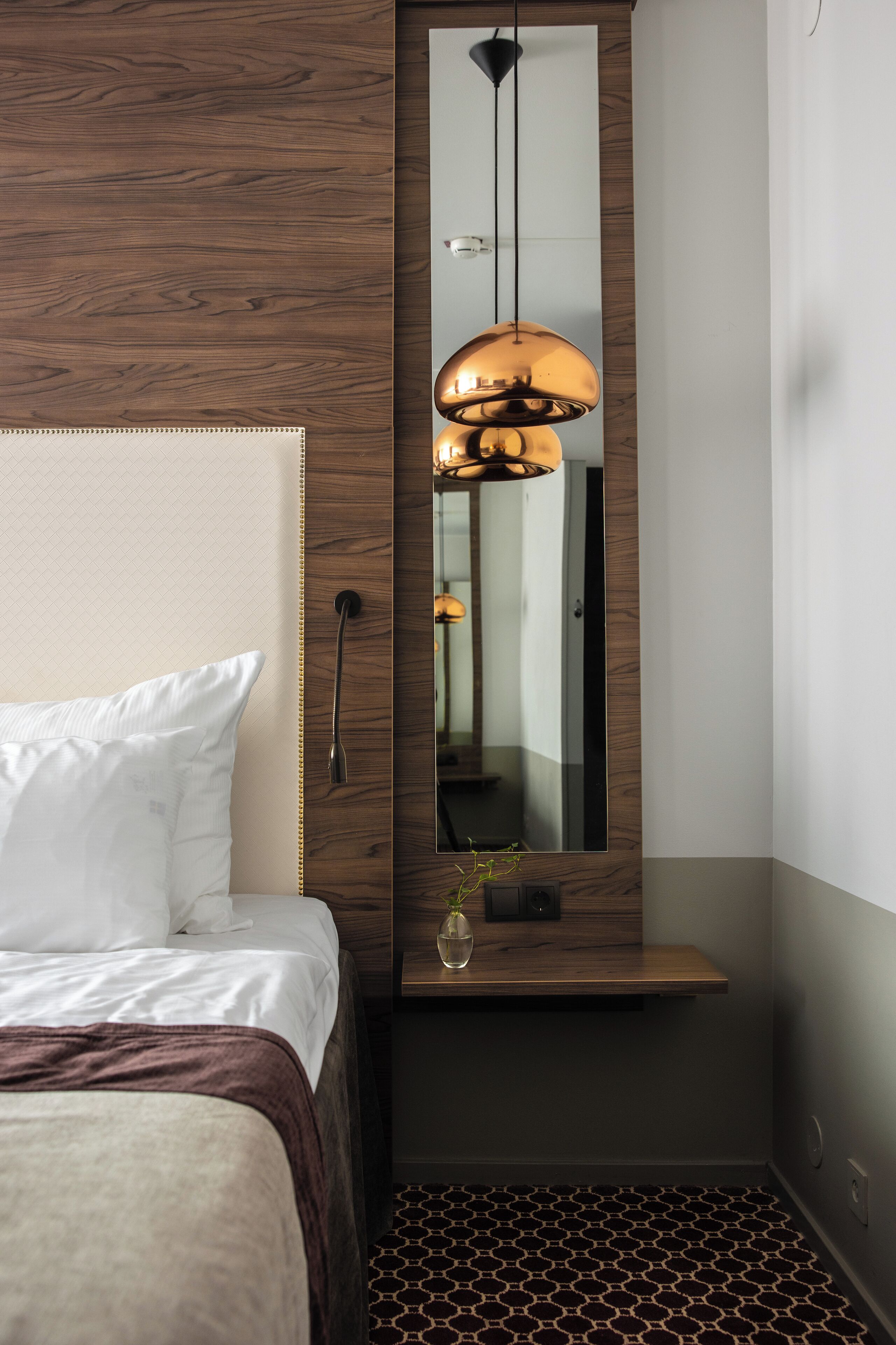 Una habitación de hotel moderna con una cama grande, cabecera de madera, una lámpara colgante, un espejo y una pequeña planta sobre una mesita de noche.