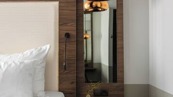 Una habitación de hotel moderna con una cama grande, cabecera de madera, una lámpara colgante, un espejo y una pequeña planta sobre una mesita de noche.