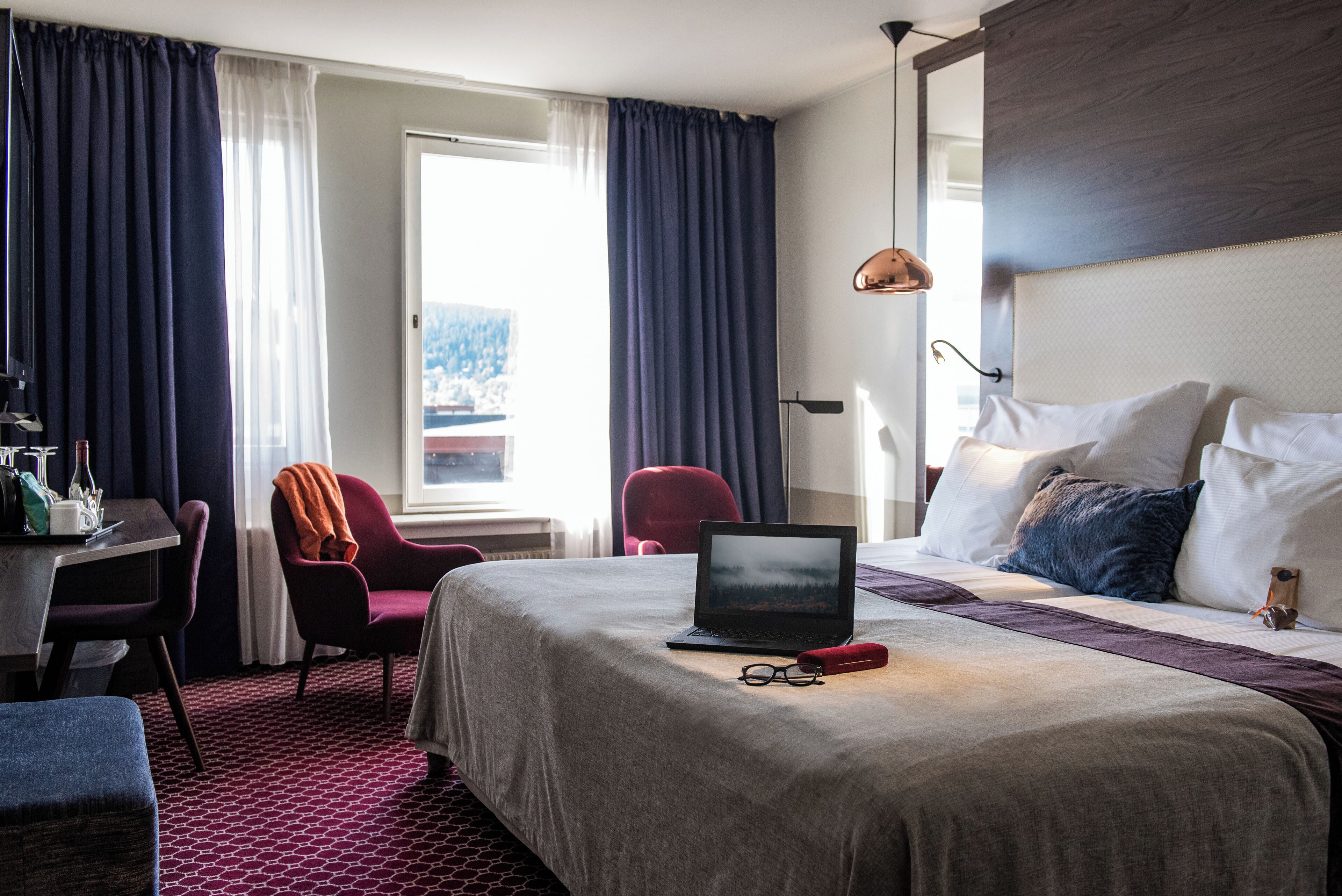 A hotel room with a bed, a desk, a chair, a laptop, and a window with curtains.