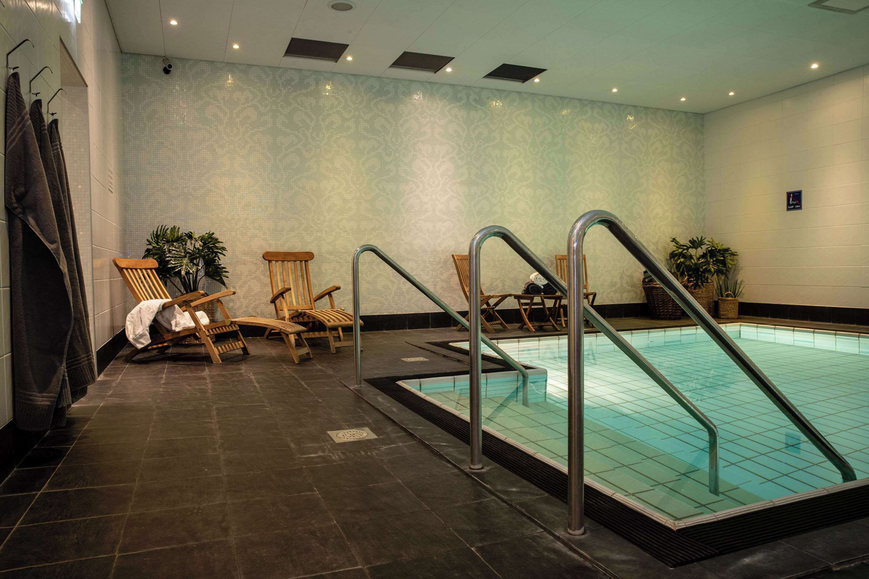 Una piscina con escaleras de metal, sillas de madera y plantas en macetas, en un espacio con paredes estampadas y suelo de baldosas oscuras.
