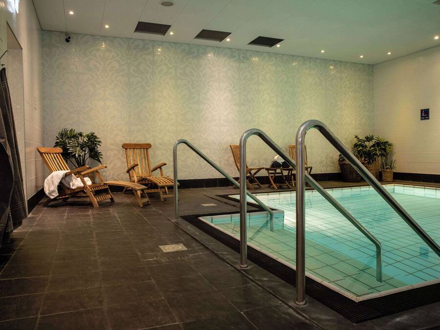 Una piscina con escaleras de metal, sillas de madera y plantas en macetas, en un espacio con paredes estampadas y suelo de baldosas oscuras.