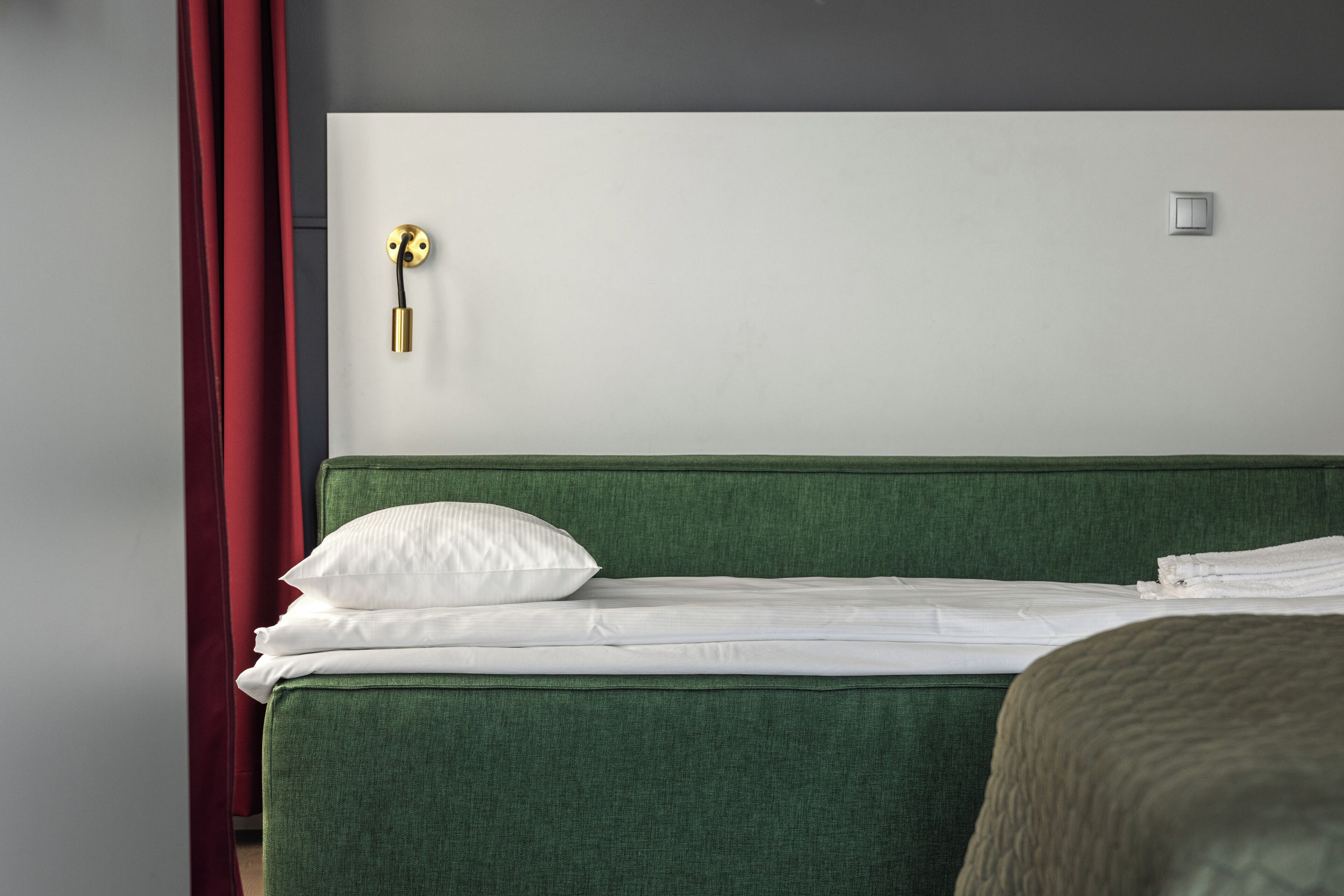 A neatly made bed with white linens and a green headboard, a wall-mounted light fixture, and a door with a golden handle.