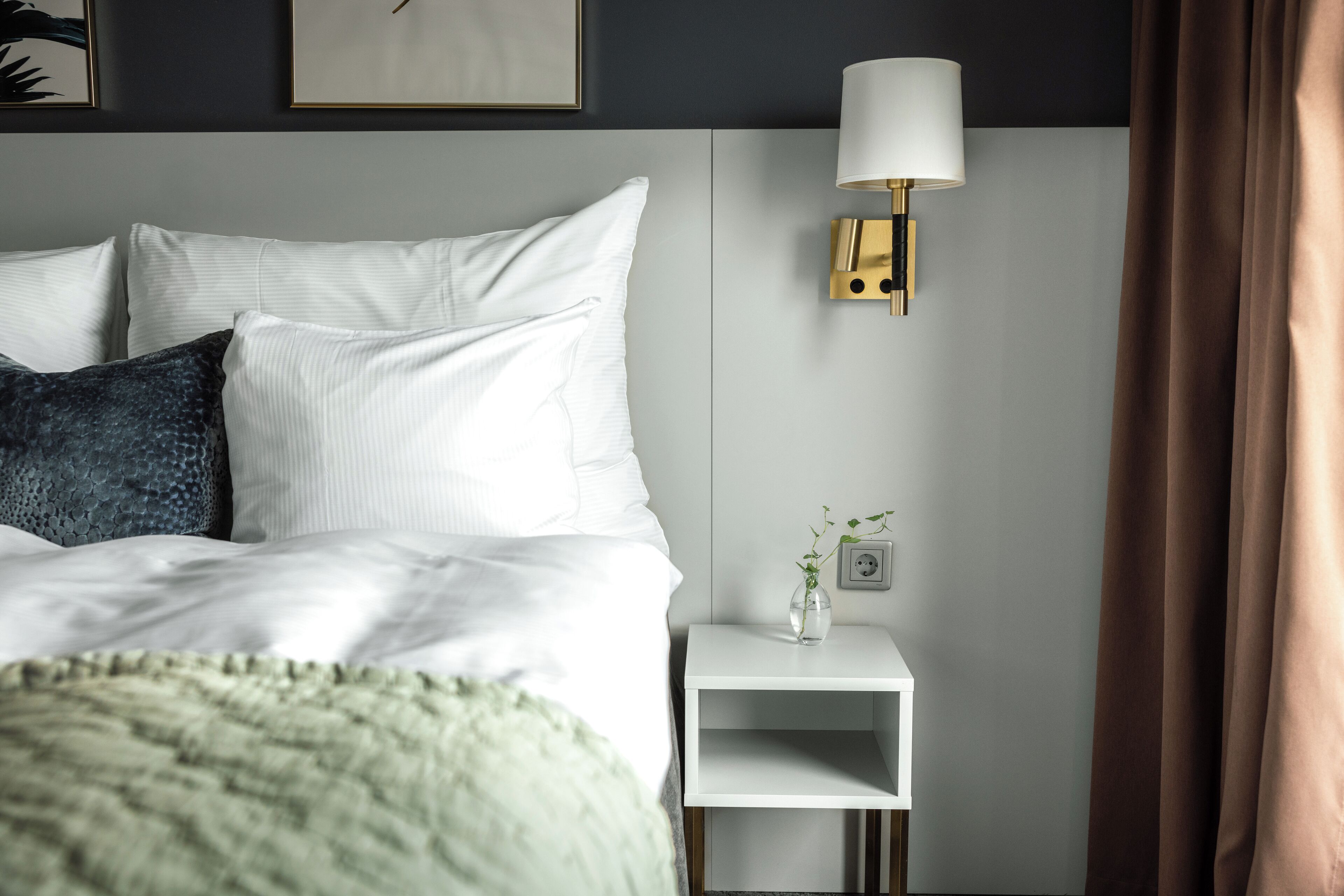 Una cama bien tendida con almohadas blancas y azul oscuro, una mesita de noche con una lámpara y un pequeño florero con una planta.