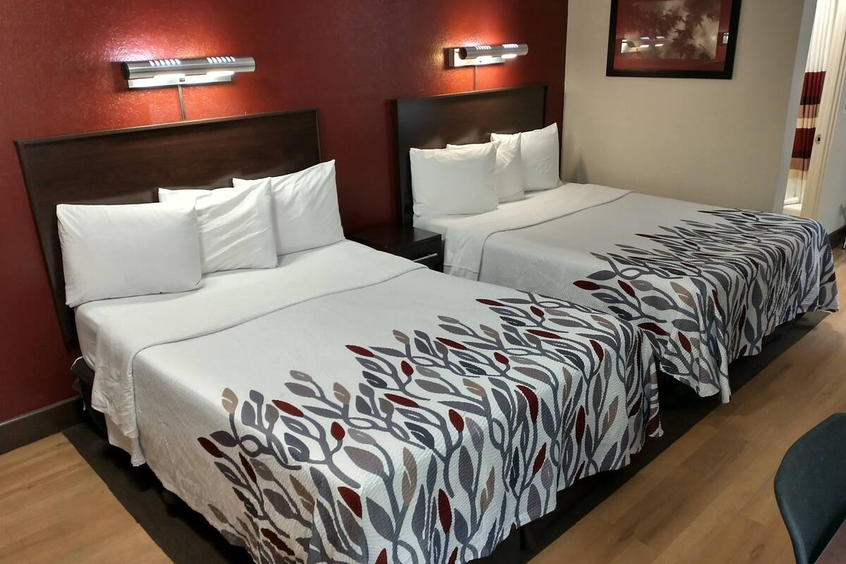 Ein Hotelzimmer mit zwei Betten, jeweils mit weißer Bettwäsche und einem gemusterten Läufer. Über jedem Bett befinden sich Wandleuchten, und an der Wand hängt ein gerahmtes Bild.