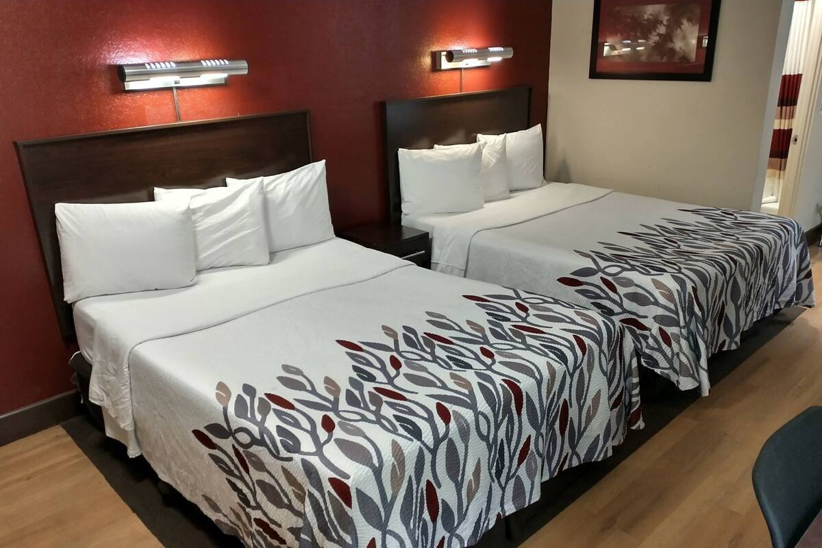 Ein Hotelzimmer mit zwei Betten, beide mit weißer Bettwäsche und einem gemusterten Läufer. Über jedem Bett befinden sich Wandleuchten.
