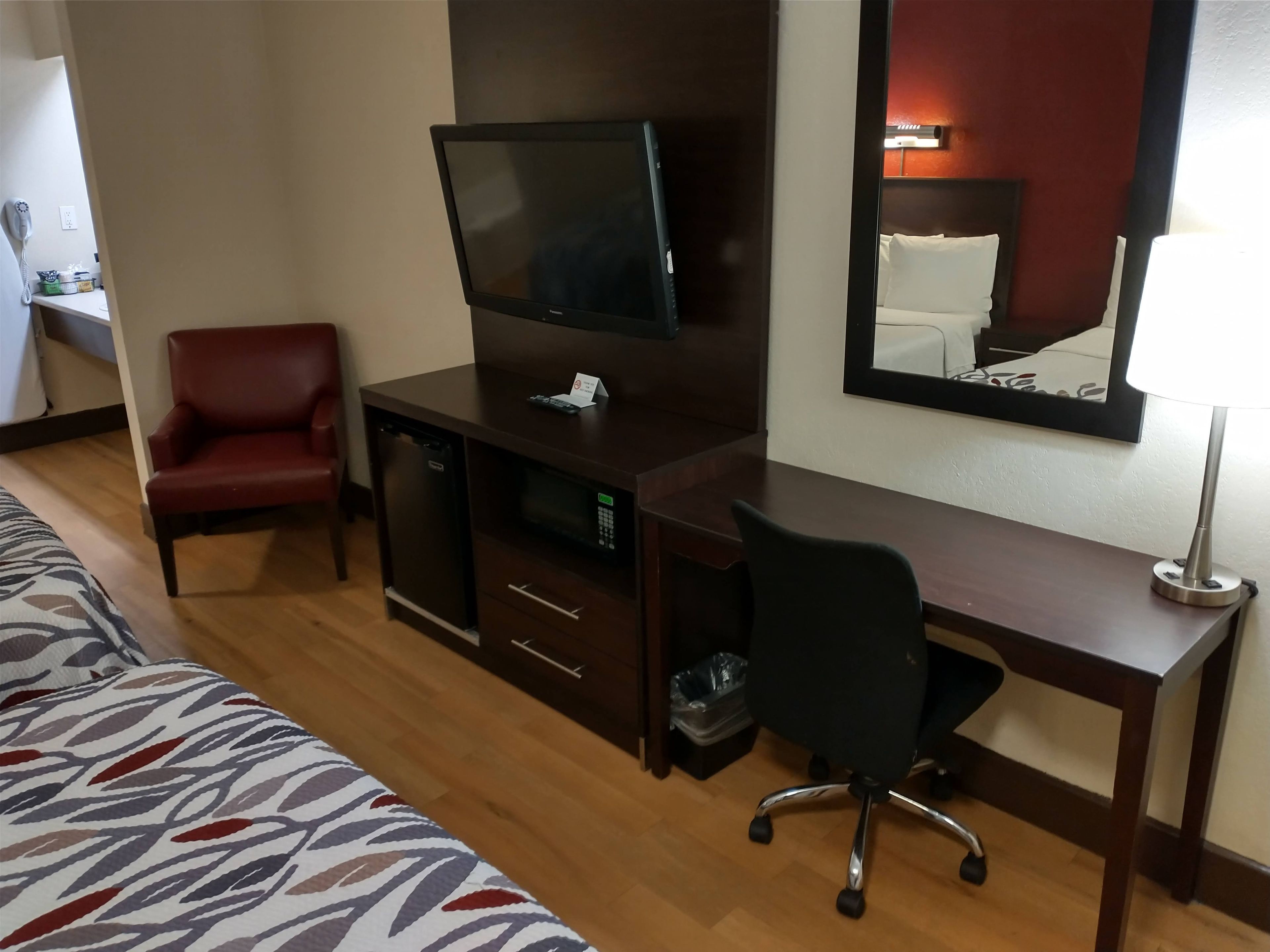 A hotel room with a bed, a desk with a television, a red chair, and a desk lamp.