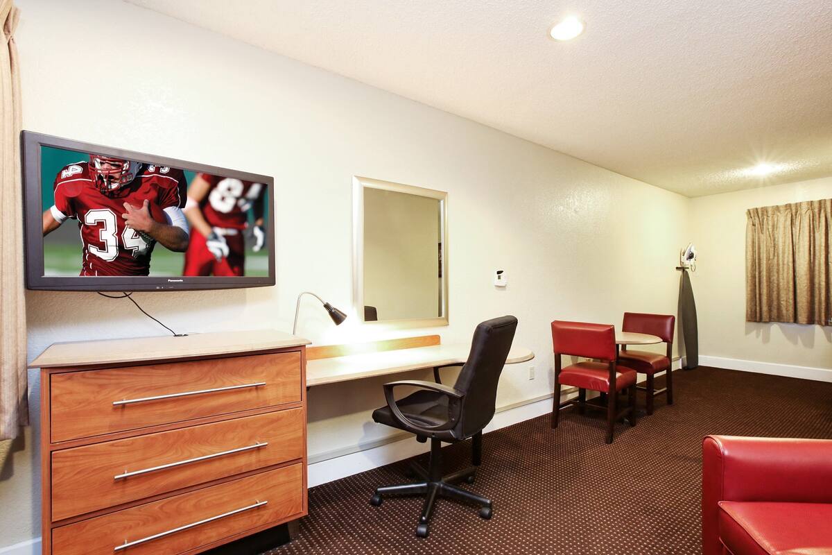 Ein Hotelzimmer mit Fernseher, der ein Sportspiel zeigt, einem Schreibtisch, einem Sessel, einer roten Couch und einem Spiegel.