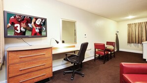 A hotel room with a television mounted on the wall, a desk with drawers, a chair, a small table, and a red couch.