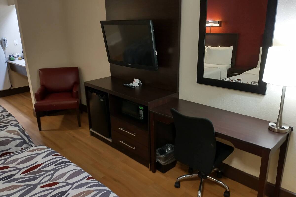 A hotel room with a bed, a desk with a computer and a flat-screen TV, a red chair, and a mirror on the wall.