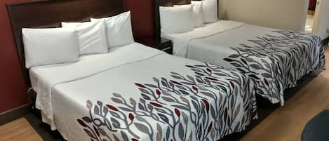 A hotel room with two beds, each with white bedding and a patterned runner. There are wall-mounted lights above each bed, and a framed picture on the wall.