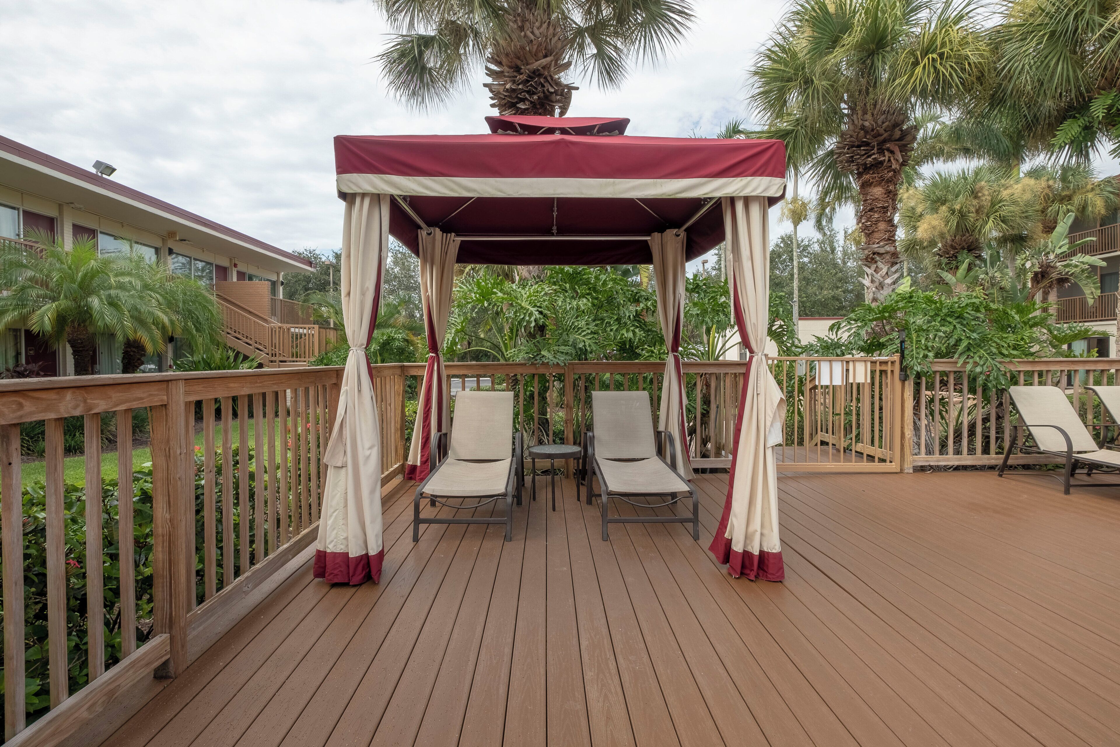 A covered outdoor seating area with a wooden deck, surrounded by a wooden railing, and featuring two lounge chairs and a small table.