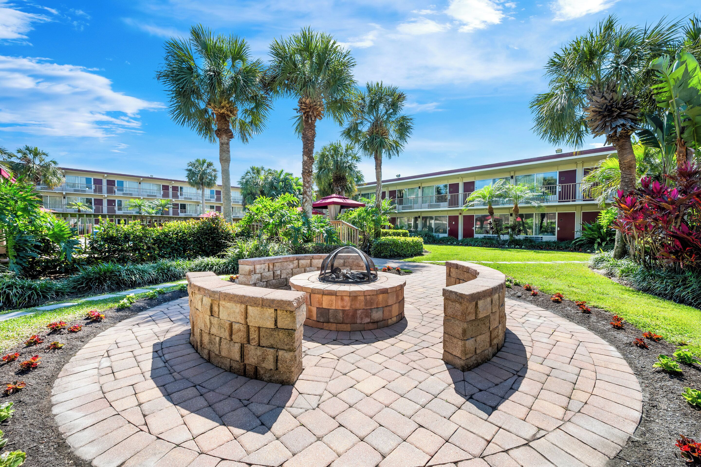 A courtyard with a circular brick firepit, palm trees, and a building with balconies in the background.