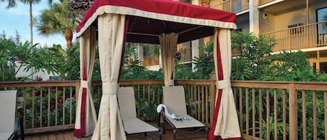 Un espace extérieur aménagé, abrité et doté de deux fauteuils et d’une table, entouré d’une terrasse en bois et de palmiers.