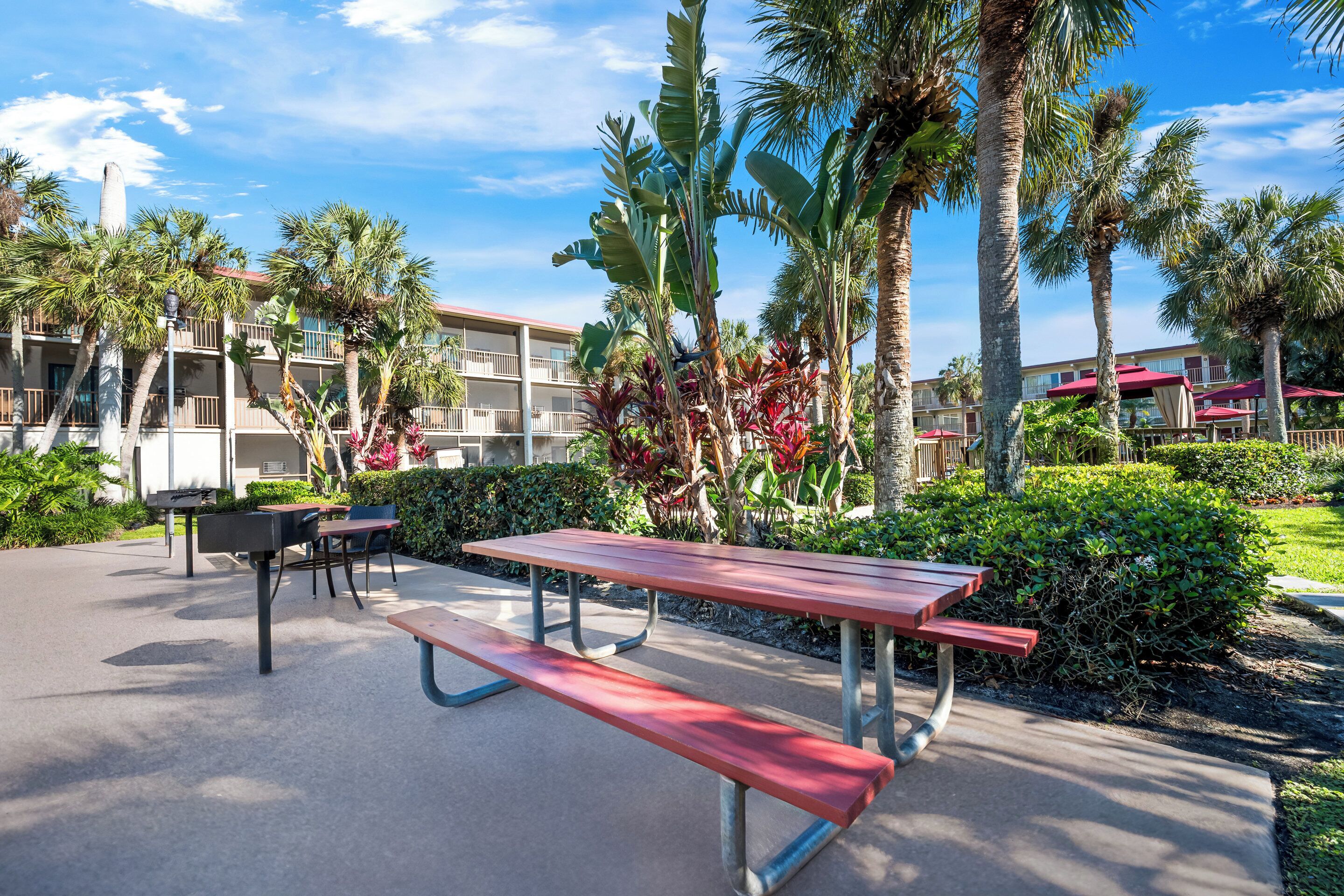 A picnic area with benches and tables, surrounded by palm trees and a building with balconies.