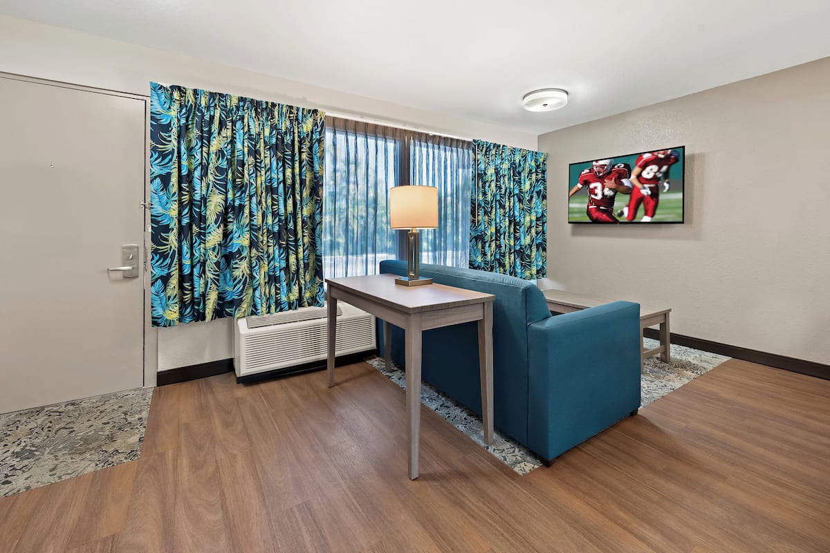 A hotel room with a blue sofa, a small table, a lamp, and a TV on the wall.