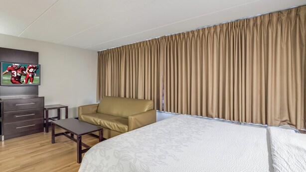 A hotel room with a bed, a sofa, a TV, a dresser, and a window with curtains. - Red Roof Inn PLUS+ Boston - Woburn/ Burlington (Woburn)