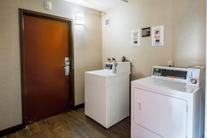A laundry room with a front-loading washer and dryer, a wooden door, and instructional posters on the wall. - Red Roof Inn PLUS+ Boston - Woburn/ Burlington (Woburn)
