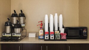 Una estación de café con máquinas de café, vasos desechables, un microondas y leche condensada.