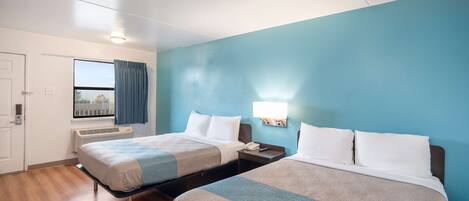 A hotel room with two beds, a window with curtains, a bedside table with a lamp, and a door.