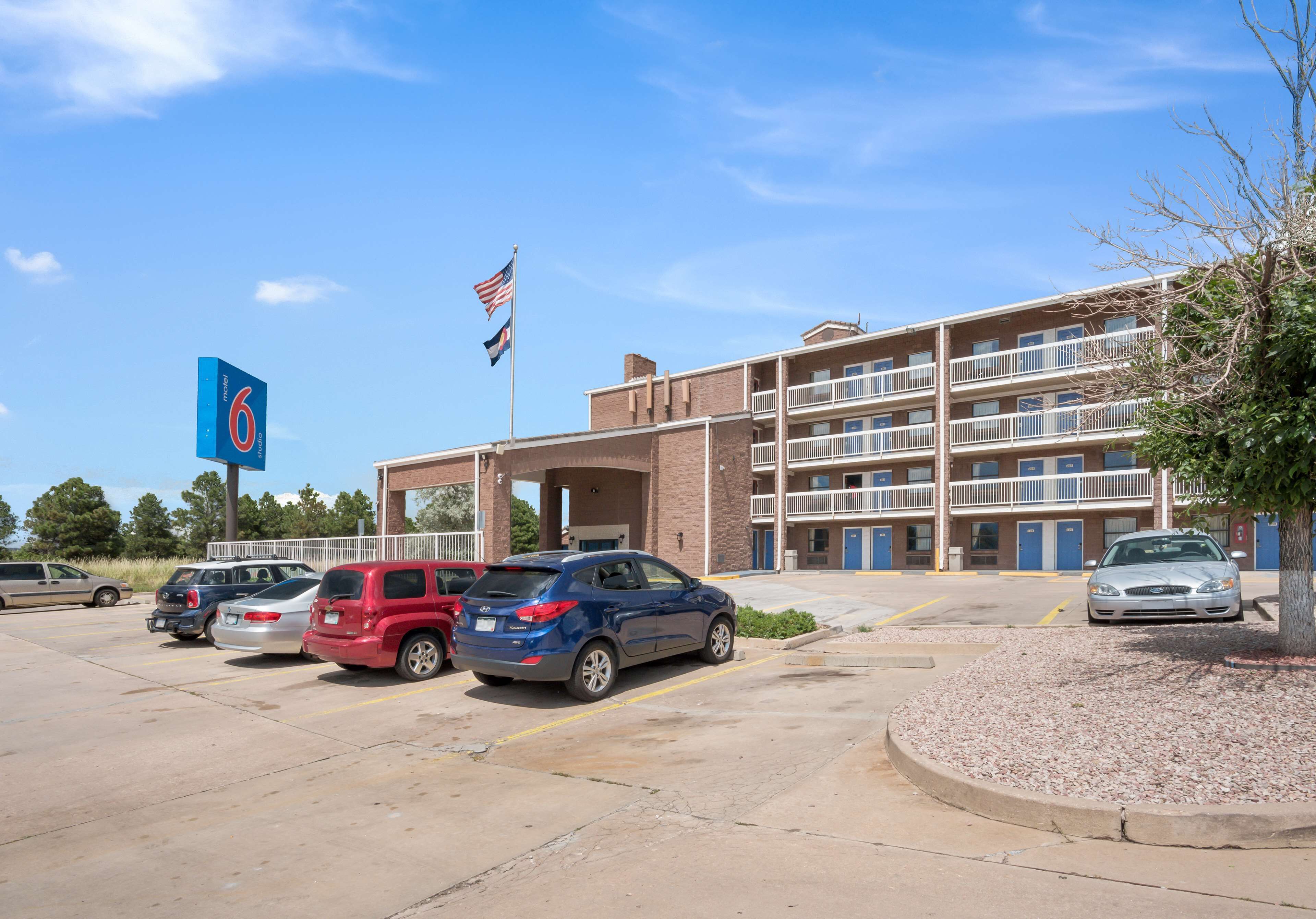 A parking lot with several cars, a building with multiple floors and balconies, and a flagpole with an American flag.