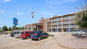 A parking lot with several cars, a building with multiple floors and balconies, and a flagpole with an American flag.