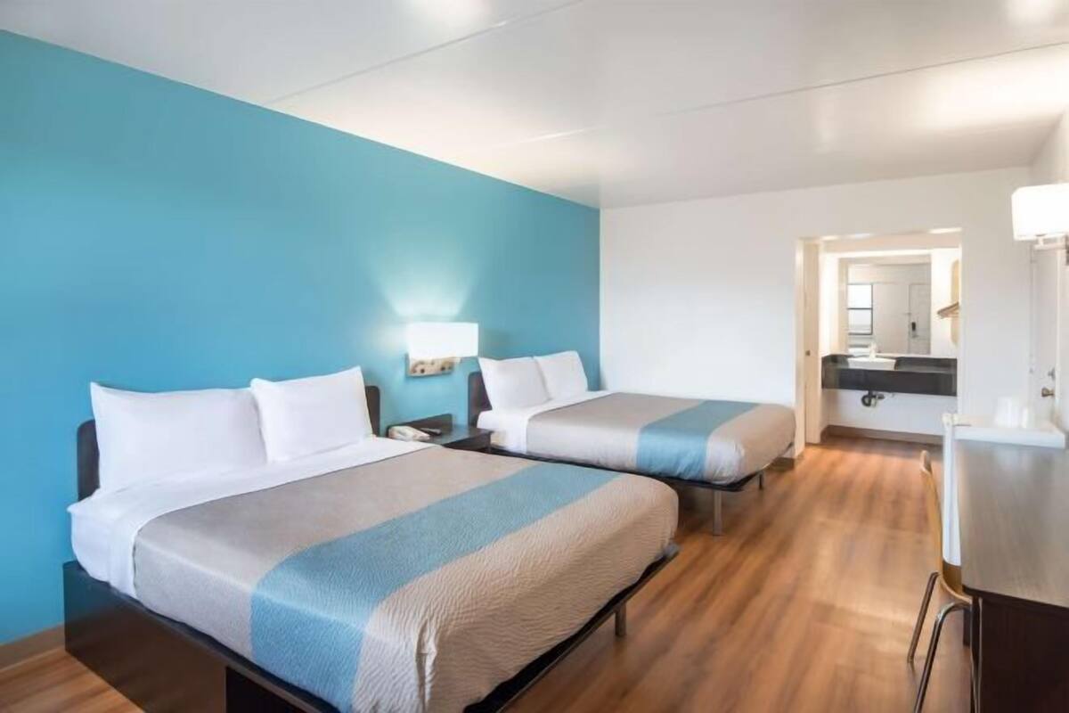 Habitación de hotel con dos camas, un escritorio, un espejo y un baño visible a través de una puerta abierta.