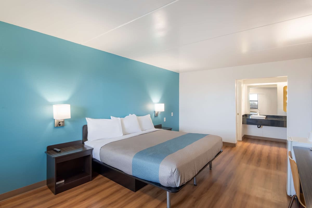 Una habitación de hotel moderna con una cama grande, mesitas de noche, un tocador, un baño con lavabo y espejo, y suelo de madera.