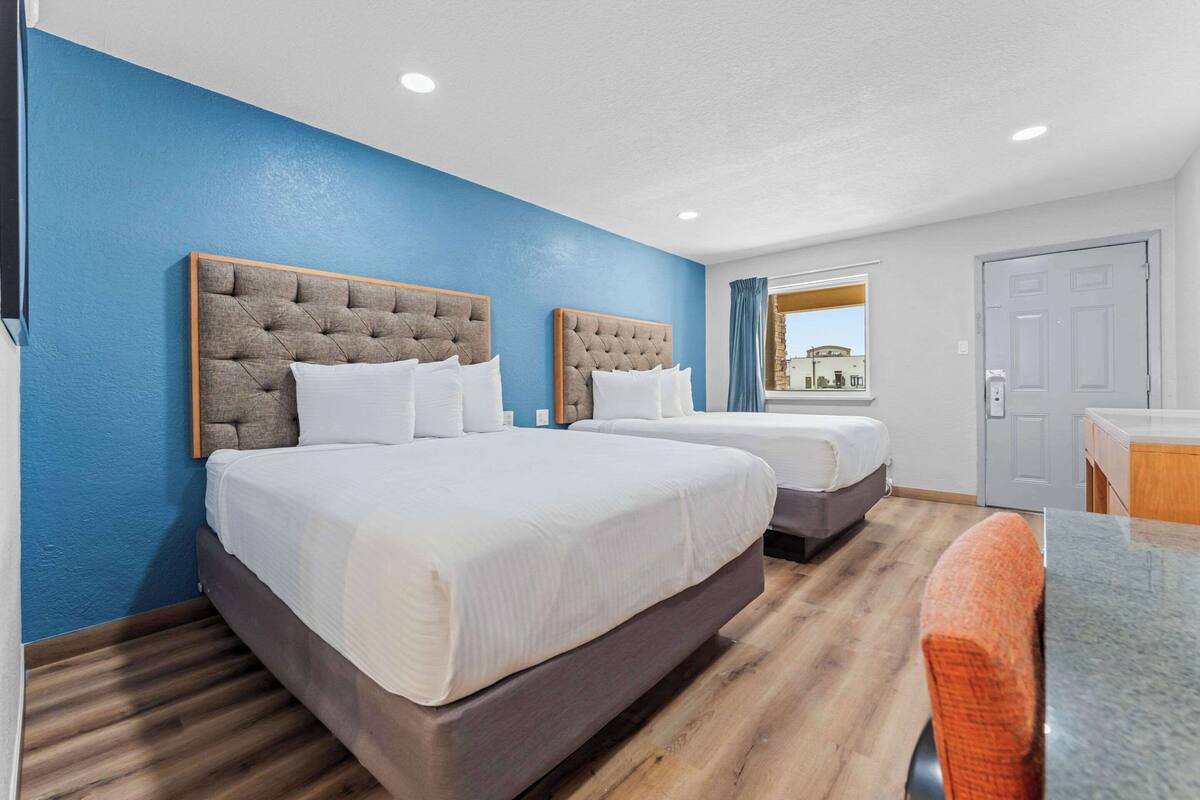 A hotel room with two beds, a wooden floor, a blue wall, and a view of the outside.