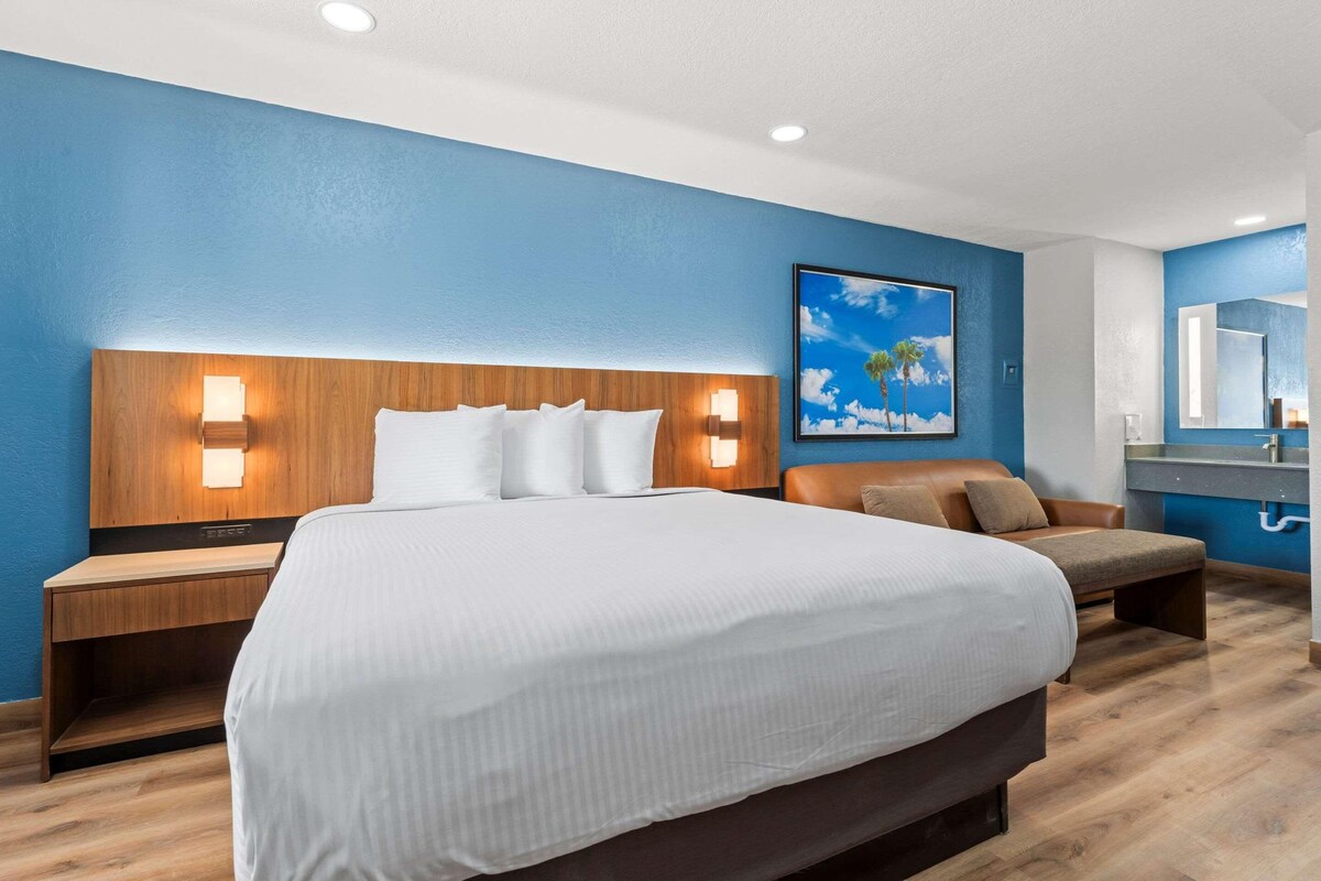 Une chambre d’hôtel avec un grand lit, une tête de lit en bois, deux tables de chevet avec des lampes, un canapé en cuir marron, un tableau au mur et une salle de bain avec un miroir et un lavabo.