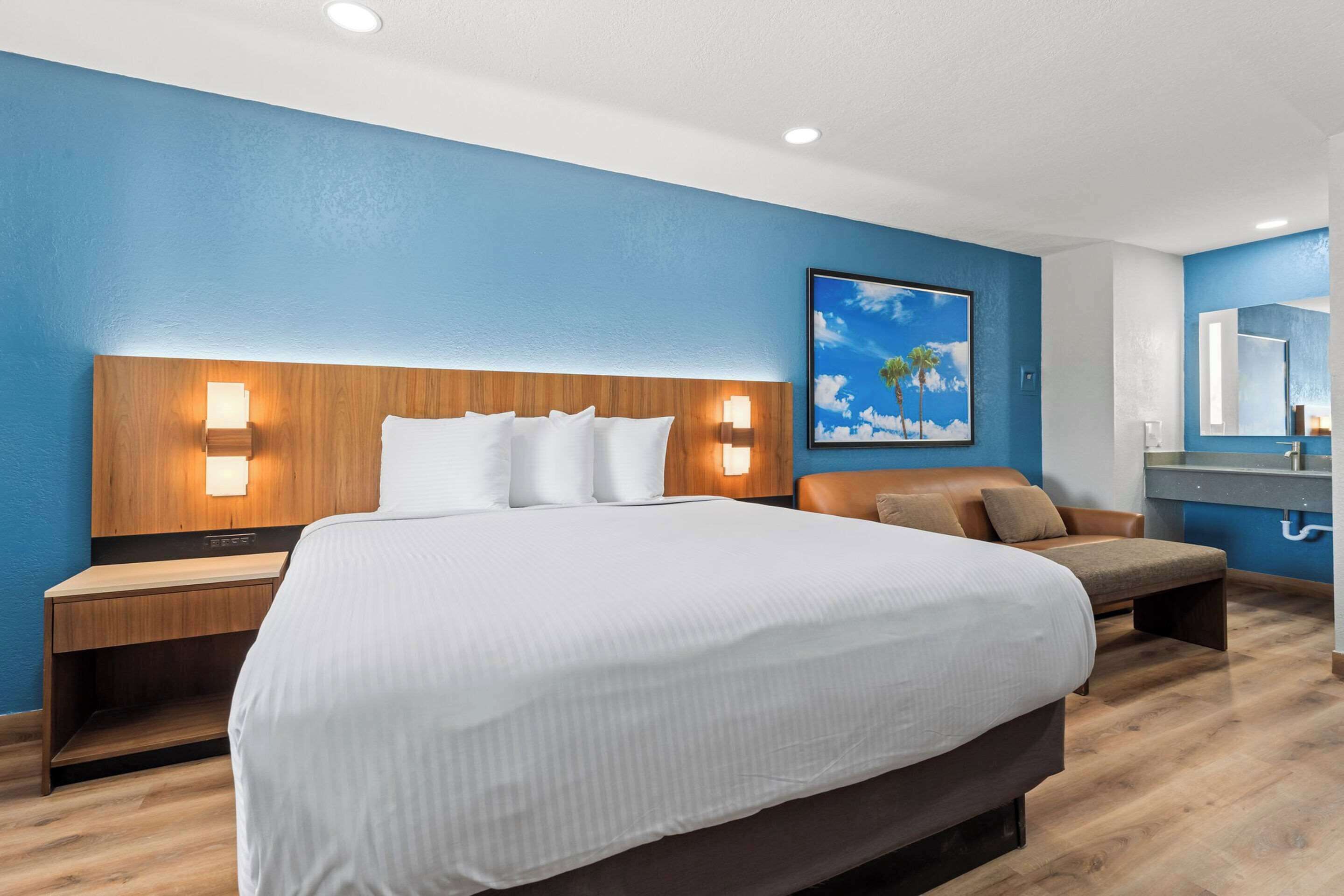 A hotel room with a large bed, wooden headboard, two bedside tables with lamps, a brown leather sofa, a framed picture on the wall, and a bathroom with a mirror and sink.
