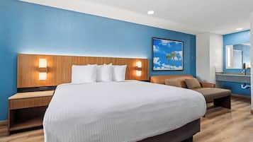 A hotel room with a large bed, wooden headboard, two bedside tables with lamps, a brown leather sofa, a framed picture on the wall, and a bathroom with a mirror and sink.