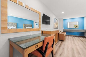Una habitación de hotel moderna con una cama grande, un escritorio con una silla, un televisor y un baño con pared azul y un espejo amplio.