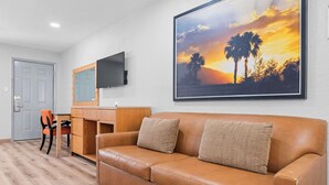A room with a brown leather sofa, a wooden desk, a television mounted on the wall, and a framed sunset painting. - Econo Lodge Inn & Suites Sealy (Sealy)