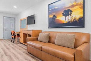 Una habitación con un sofá de cuero marrón, un escritorio de madera, un televisor montado en la pared y un cuadro con una vista de atardecer enmarcado.