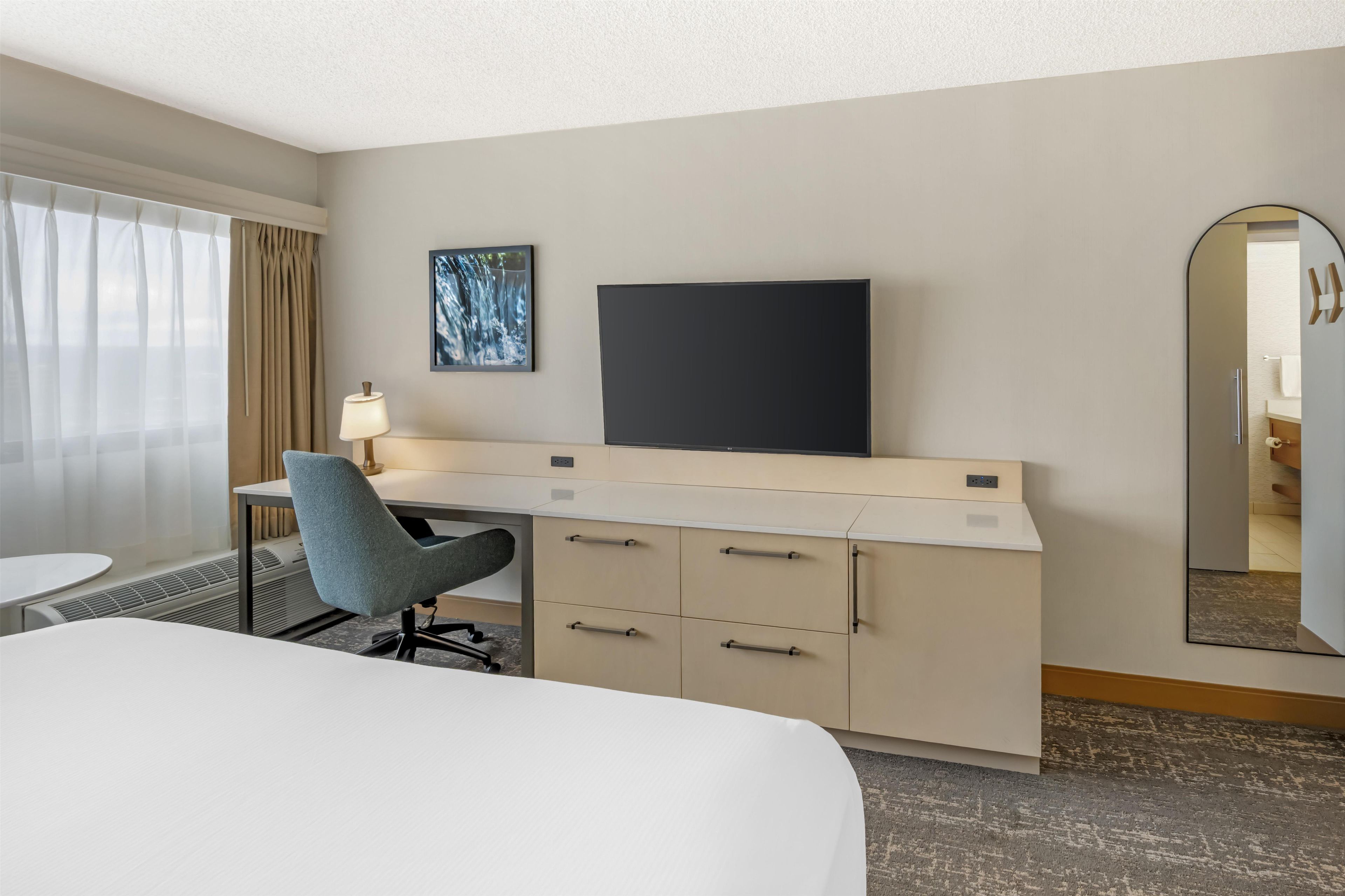 A hotel room with a bed, a desk with a chair, a flat-screen TV, a painting, and a mirror.