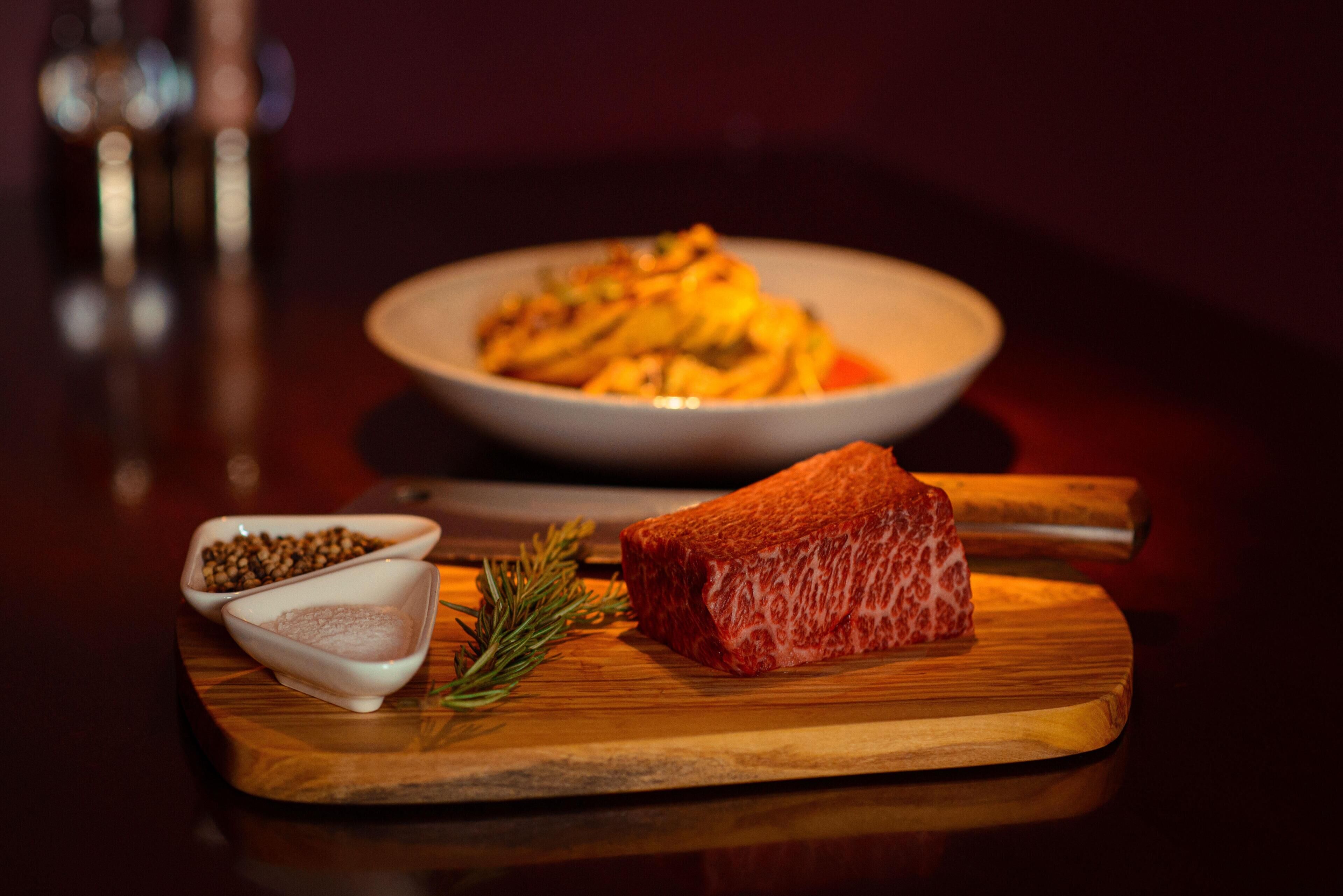 A wooden cutting board with a piece of meat and two small bowls containing spices and herbs.
