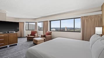 Habitación de hotel con una cama grande, televisor, escritorio y una silla. Hay ventanas con cortinas y vistas a edificios.