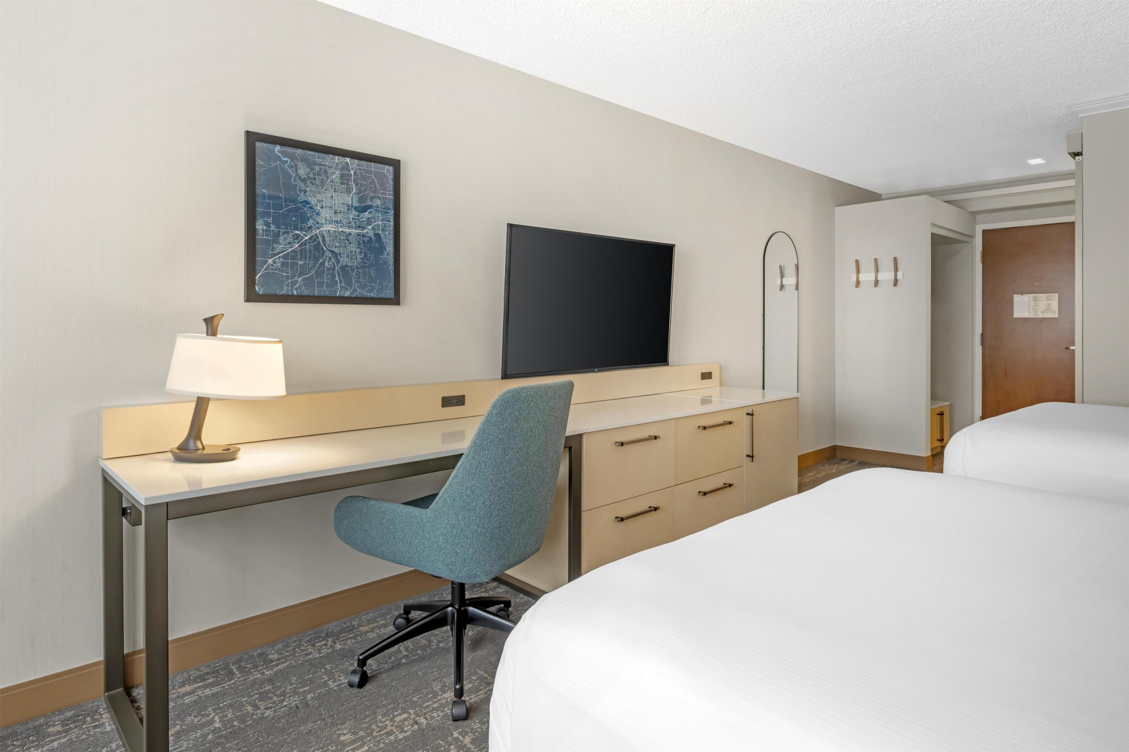 A hotel room with a desk, chair, TV, and two beds.