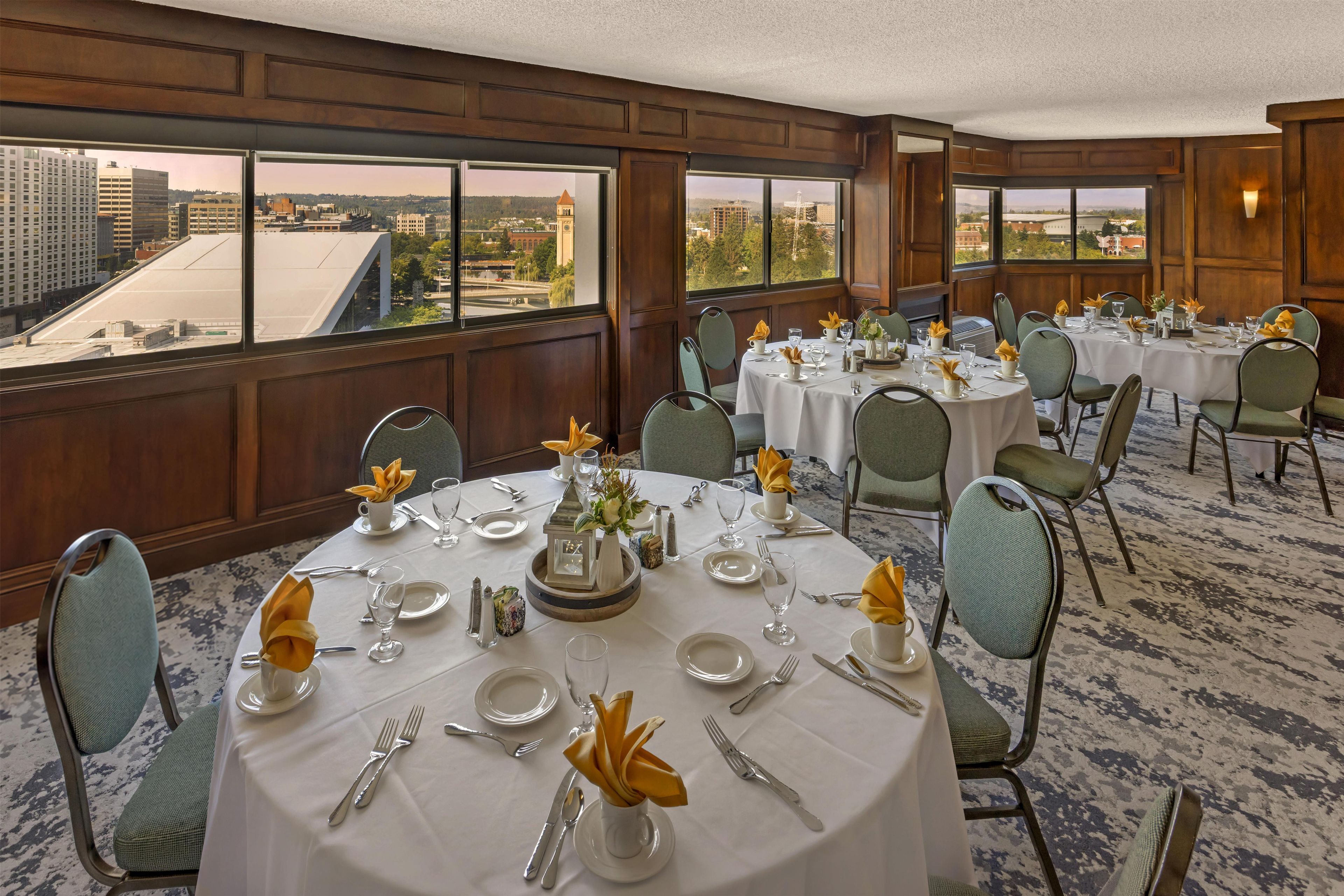 A hotel dining room with round tables set for a meal, large windows offering a city view, and a carpeted floor.