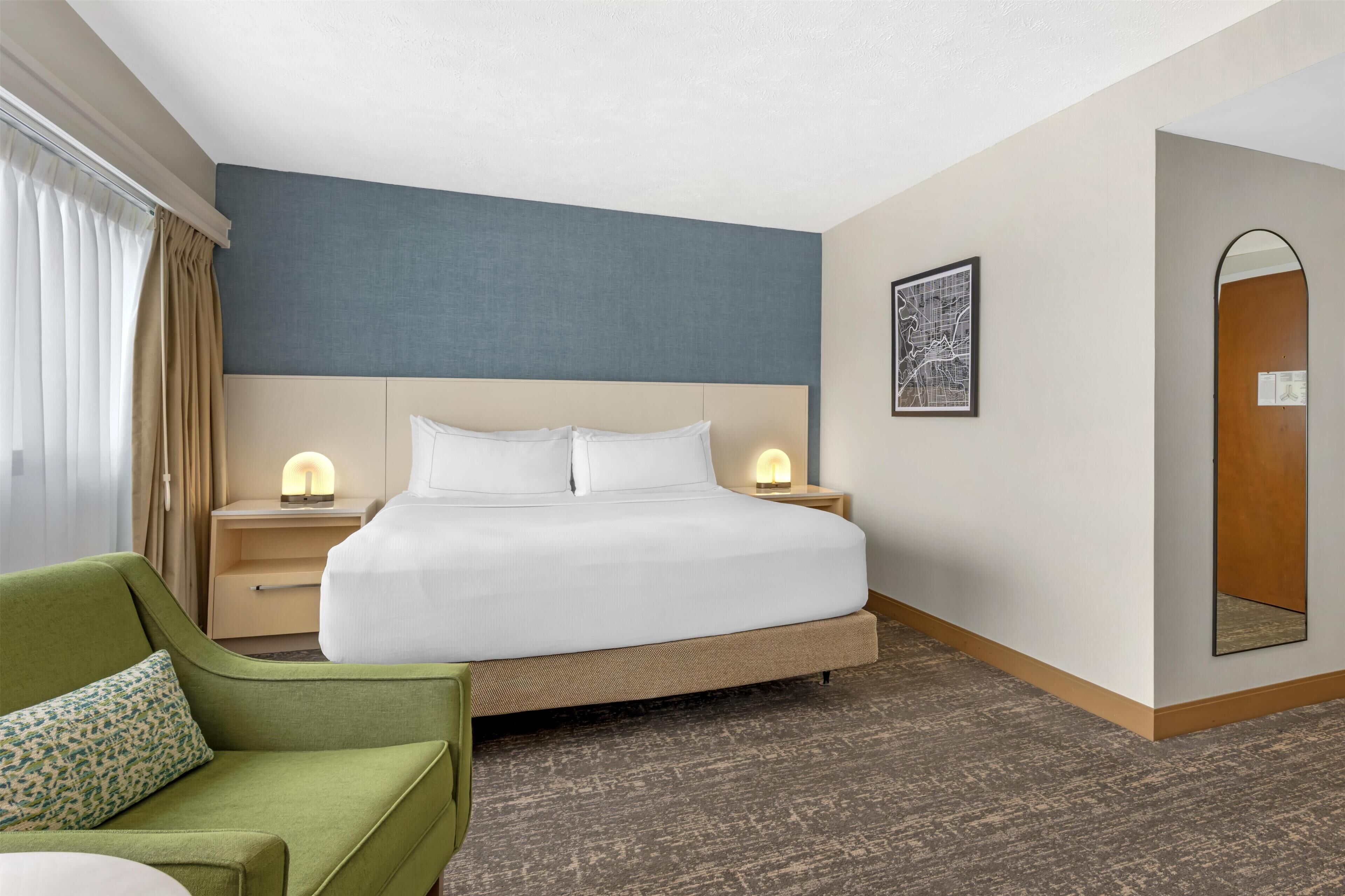 A hotel room with a bed, nightstands, a green armchair, a mirror, and a wall with a framed picture.