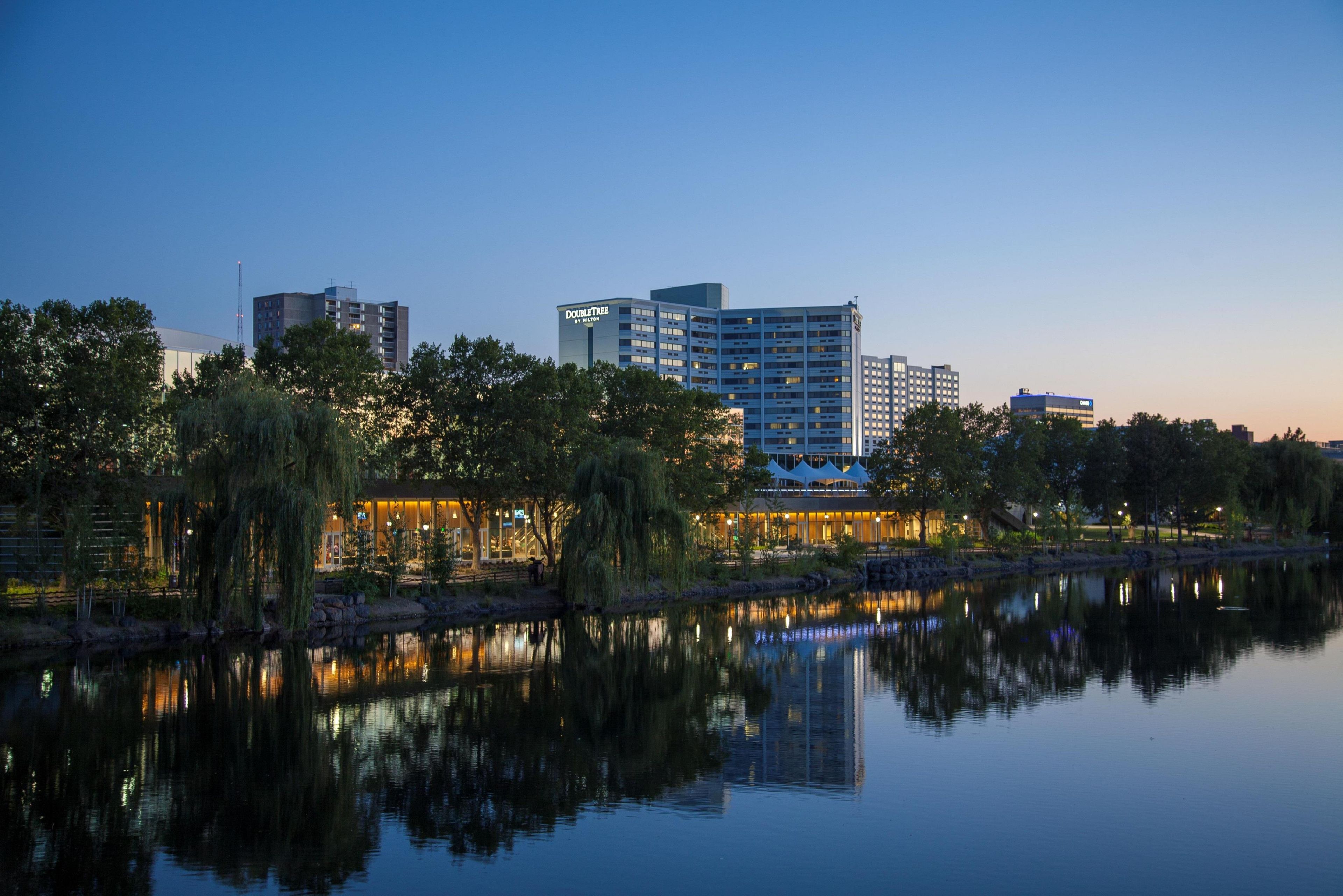 A hotel by the water with a reflection in the lake, surrounded by trees and other buildings.
