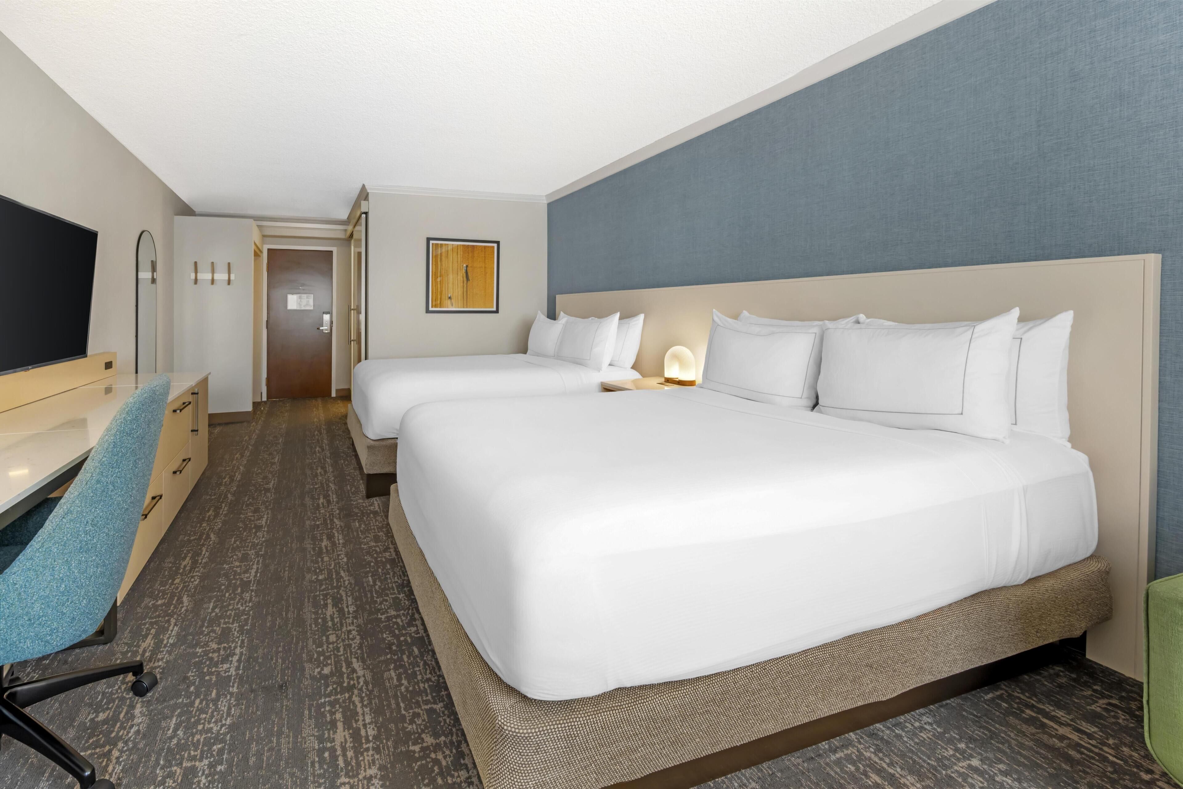 Habitación de hotel con dos camas, un escritorio con silla, un televisor y una puerta que da a otra habitación.