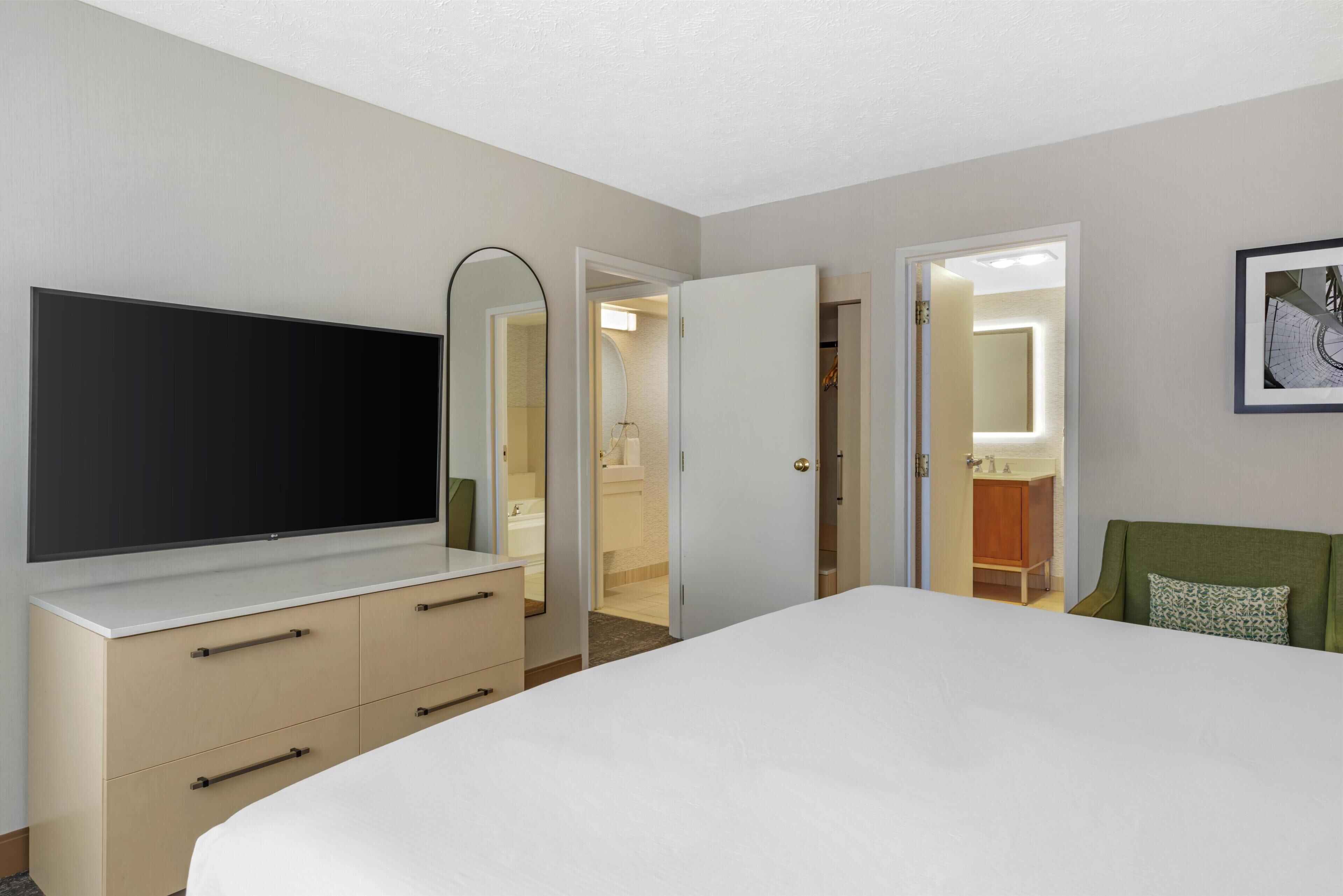 A hotel room with a bed, a TV, a dresser, a mirror, and an open door leading to a bathroom.