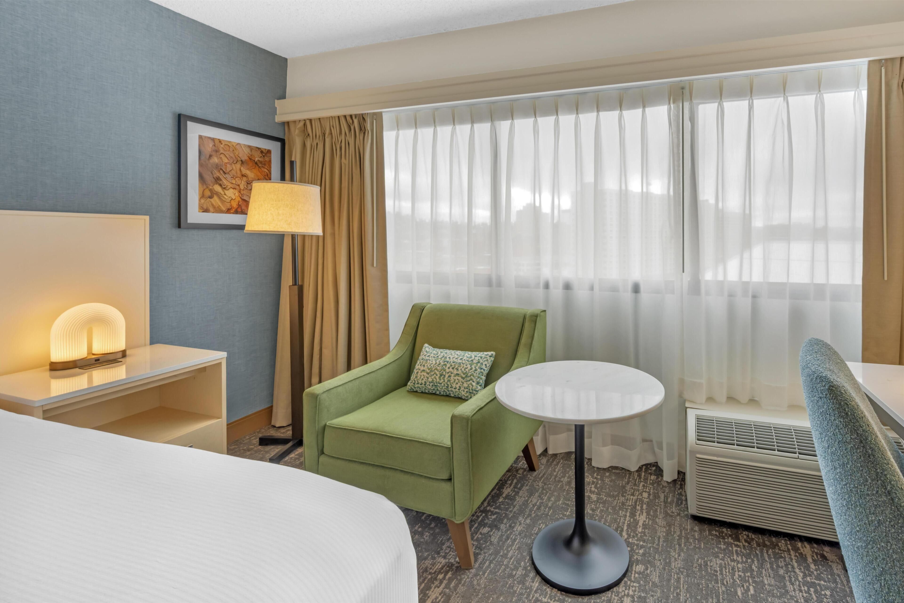 A hotel room with a bed, a green armchair, a round table, a lamp, a picture on the wall, and a large window with curtains.