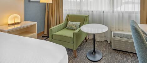 A hotel room with a bed, a green armchair, a round table, a lamp, a picture on the wall, and a large window with curtains.