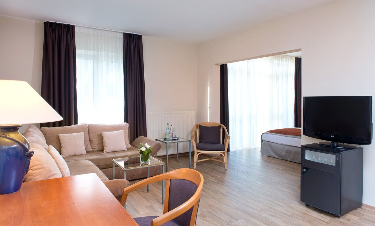 A modern hotel room with a sofa, armchair, TV, and a small table with a lamp.