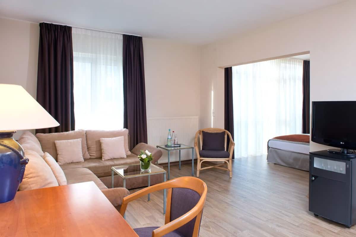 Moderni hotellihuone, jossa on sohva, nojatuoli, televisio ja pieni pöytä, jossa on lamppu.