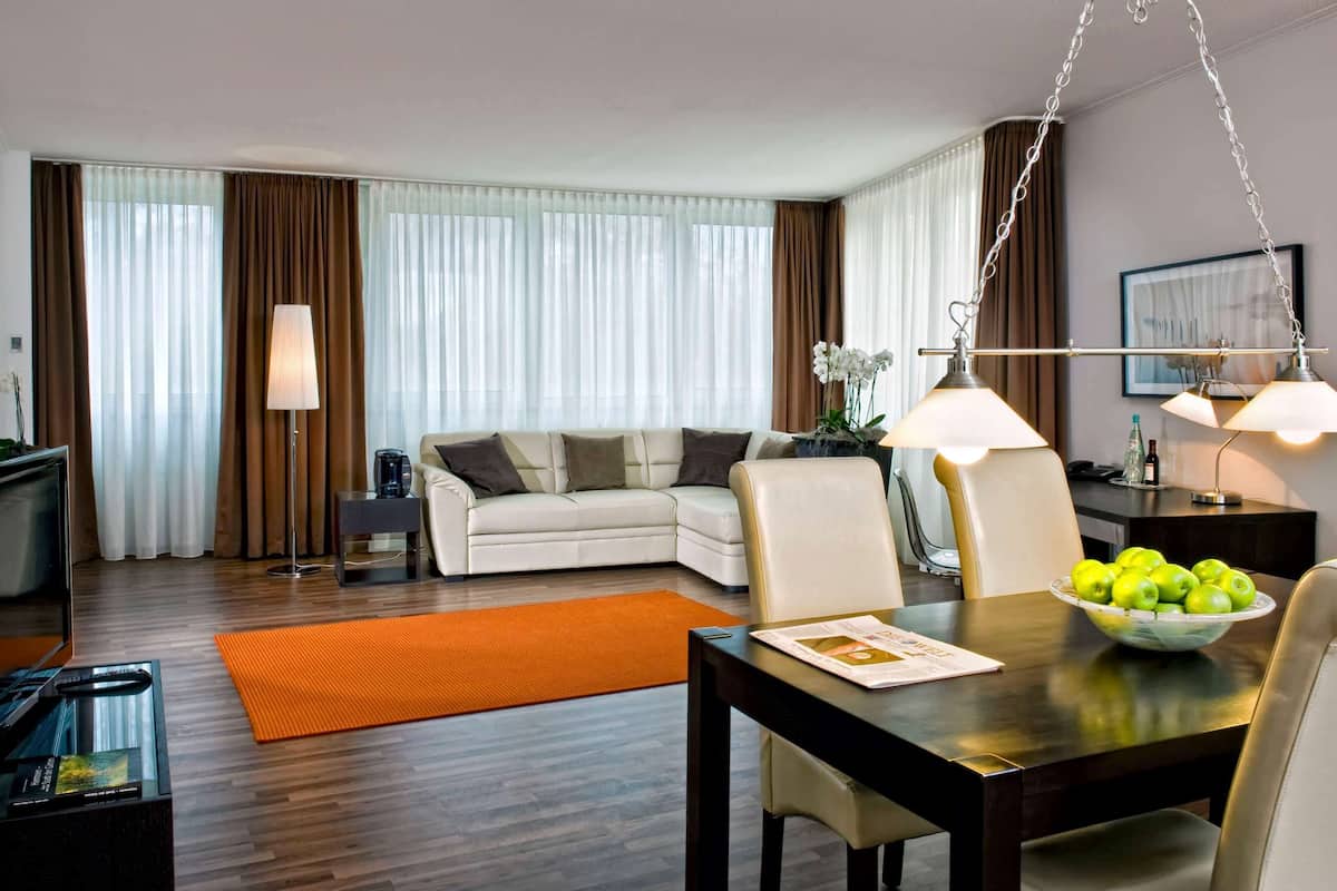 A modern living room with a sofa, a dining table, and a TV. There is a coffee table with a bowl of fruit and a magazine. The room has large windows with curtains.