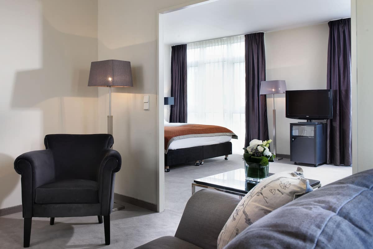 A hotel room with a black armchair, a sofa, a bed with a white mattress and brown blanket, a television, a coffee machine, and a small table with a vase of flowers.